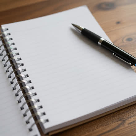 Caderno aberto com uma caneta apoiada, sobre mesa de madeira.